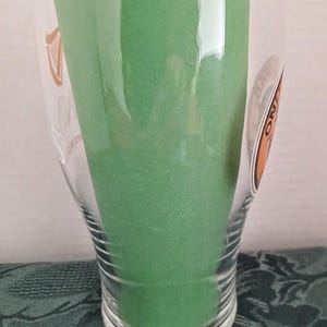 Może przedstawiać: Przezroczysty szklany kufel z logo Guinness i zielonym przedmiotem w środku. Szkło ma zakrzywiony kształt i szeroki otw&oacute;r. Zielony przedmiot wydaje się być pianką lub gąbką.