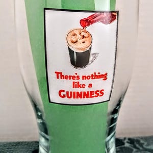 Może przedstawiać: Przezroczysty szklany kufel z zieloną wkładką. Na szkle grafika przedstawiająca nalewane piwo Guinness z czerwonym napisem "There's nothing like a GUINNESS".