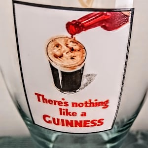 Może przedstawiać: Przezroczysty szklany kufel z nadrukowaną etykietą przedstawiającą grafikę wlewania piwa Guinness. Etykieta głosi &bdquo;There's nothing like a GUINNESS&rdquo; w czerwonym tekście. Szklanka jest częściowo wypełniona.