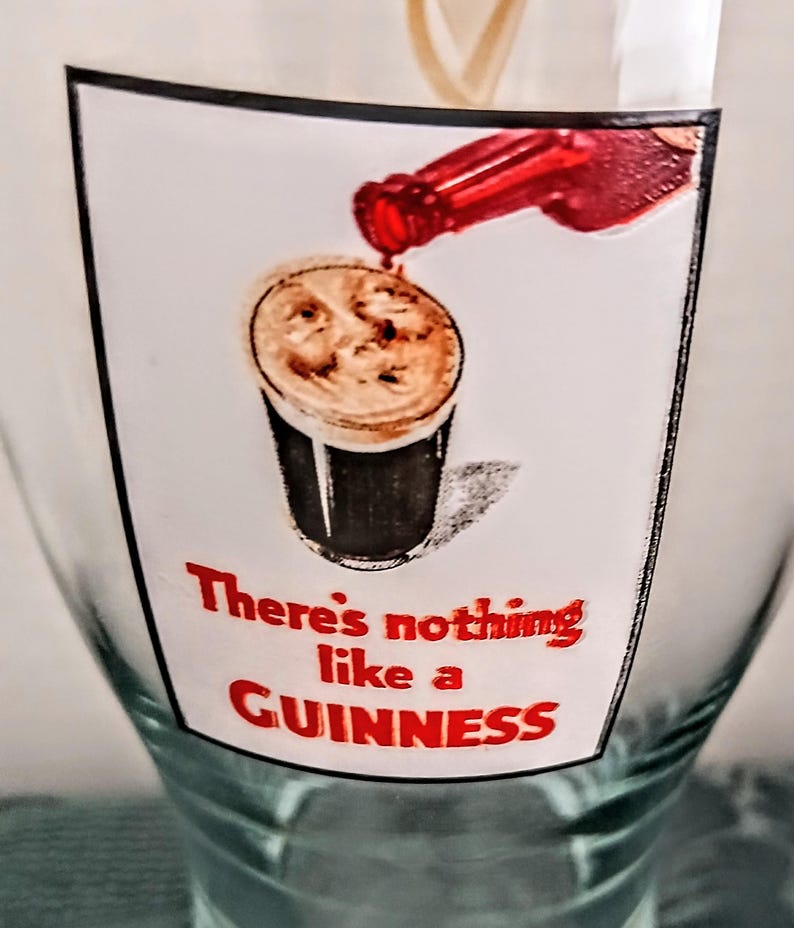 Może przedstawiać: Przezroczysty szklany kufel z nadrukowaną etykietą przedstawiającą grafikę wlewania piwa Guinness. Etykieta głosi &bdquo;There's nothing like a GUINNESS&rdquo; w czerwonym tekście. Szklanka jest częściowo wypełniona.