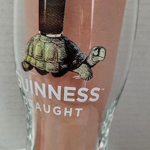 May include: A clear Guinness pint glass with a cartoon image of a turtle carrying a pint of stout. The Guinness logo is printed on the glass. The glass has a wide base and tapers upwards.