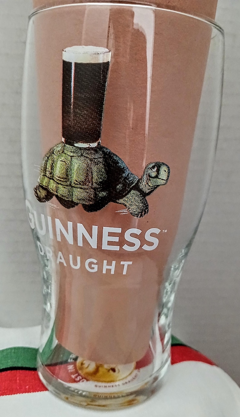 May include: A clear Guinness pint glass with a cartoon image of a turtle carrying a pint of stout. The Guinness logo is printed on the glass. The glass has a wide base and tapers upwards.
