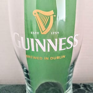 Może przedstawiać: Przezroczysty szklany kufel z logo Guinness, ze złotą harfą i napisem "GUINNESS" w kolorze białym. Widoczny jest r&oacute;wnież napis "BREWED IN DUBLIN". Szklanka jest częściowo wypełniona zieloną wkładką.