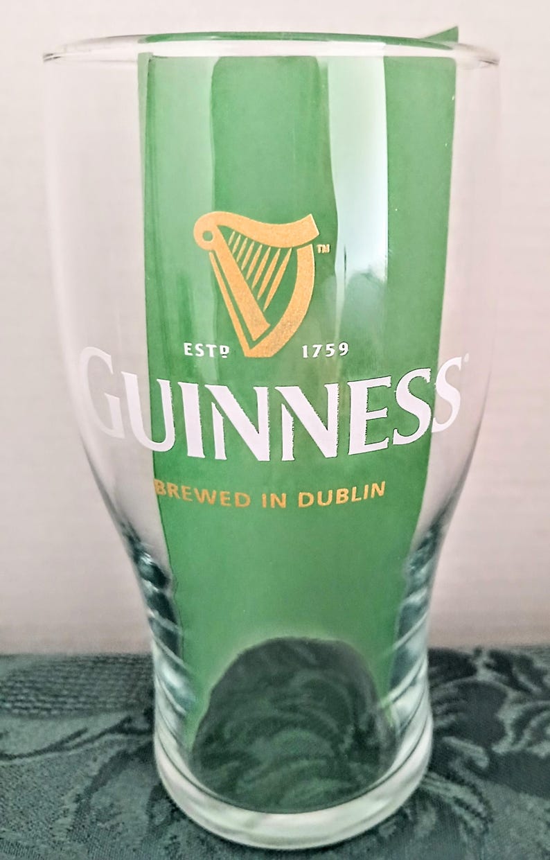 Może przedstawiać: Przezroczysty szklany kufel z logo Guinness, ze złotą harfą i napisem "GUINNESS" w kolorze białym. Widoczny jest r&oacute;wnież napis "BREWED IN DUBLIN". Szklanka jest częściowo wypełniona zieloną wkładką.