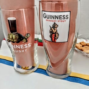 May include: Two clear Guinness pint glasses with the Guinness logo. One glass features a turtle with a pint of stout on its back, the other a bear holding a keg. Both glasses have the words "Guinness Draught Stout" printed on them.