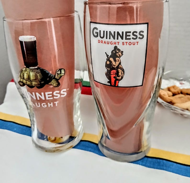 May include: Two clear Guinness pint glasses with the Guinness logo. One glass features a turtle with a pint of stout on its back, the other a bear holding a keg. Both glasses have the words "Guinness Draught Stout" printed on them.