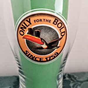 Może przedstawiać: Przezroczysty szklany kieliszek z zielonym płynem w środku, z okrągłą etykietą z tukanem i napisem "ONLY FOR THE BOLD SINCE 1759". Kieliszek ma rozszerzający się kształt.