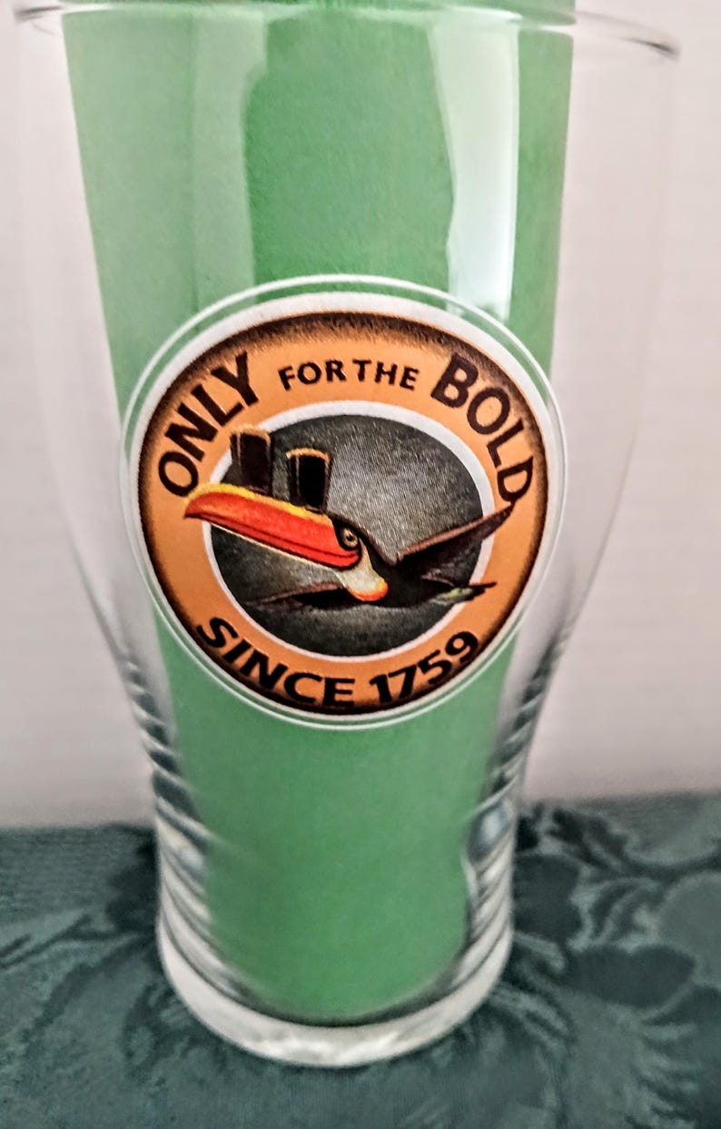 Może przedstawiać: Przezroczysty szklany kieliszek z zielonym płynem w środku, z okrągłą etykietą z tukanem i napisem "ONLY FOR THE BOLD SINCE 1759". Kieliszek ma rozszerzający się kształt.