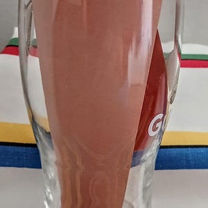 May include: A clear glass beer mug with a curved shape, holding a folded brown paper. The letter "G" is visible on the side of the glass. The background is a colorful striped fabric.