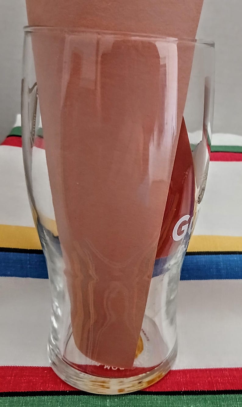 May include: A clear glass beer mug with a curved shape, holding a folded brown paper. The letter "G" is visible on the side of the glass. The background is a colorful striped fabric.