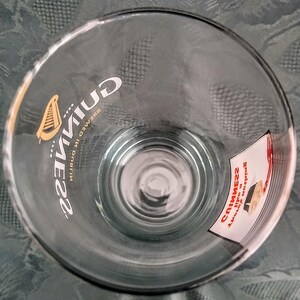 Może przedstawiać: Przezroczysty szklany kufel z logo Guinness i symbolem harfy. Widoczny jest napis "Guinness, brewed in Dublin". Do szkła przymocowana jest mała naklejka z dodatkowym tekstem.