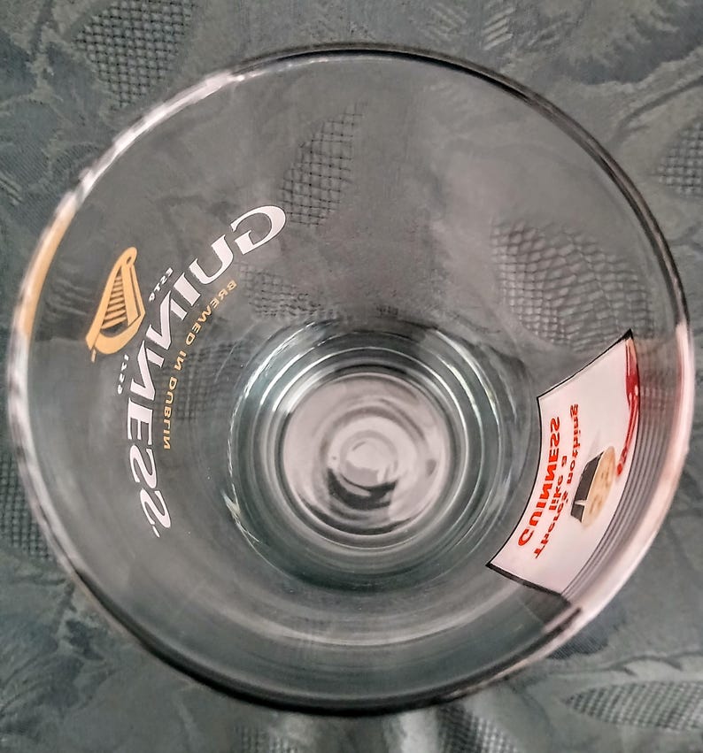 Może przedstawiać: Przezroczysty szklany kufel z logo Guinness i symbolem harfy. Widoczny jest napis "Guinness, brewed in Dublin". Do szkła przymocowana jest mała naklejka z dodatkowym tekstem.