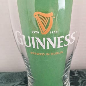 Może przedstawiać: Przezroczysty szklany kufel Guinness z białym logo Guinness i pomarańczowym symbolem harfy. Widoczny jest r&oacute;wnież tekst "ESTD 1759" i "BREWED IN DUBLIN". Szklanka wypełniona jest zielonym papierem.