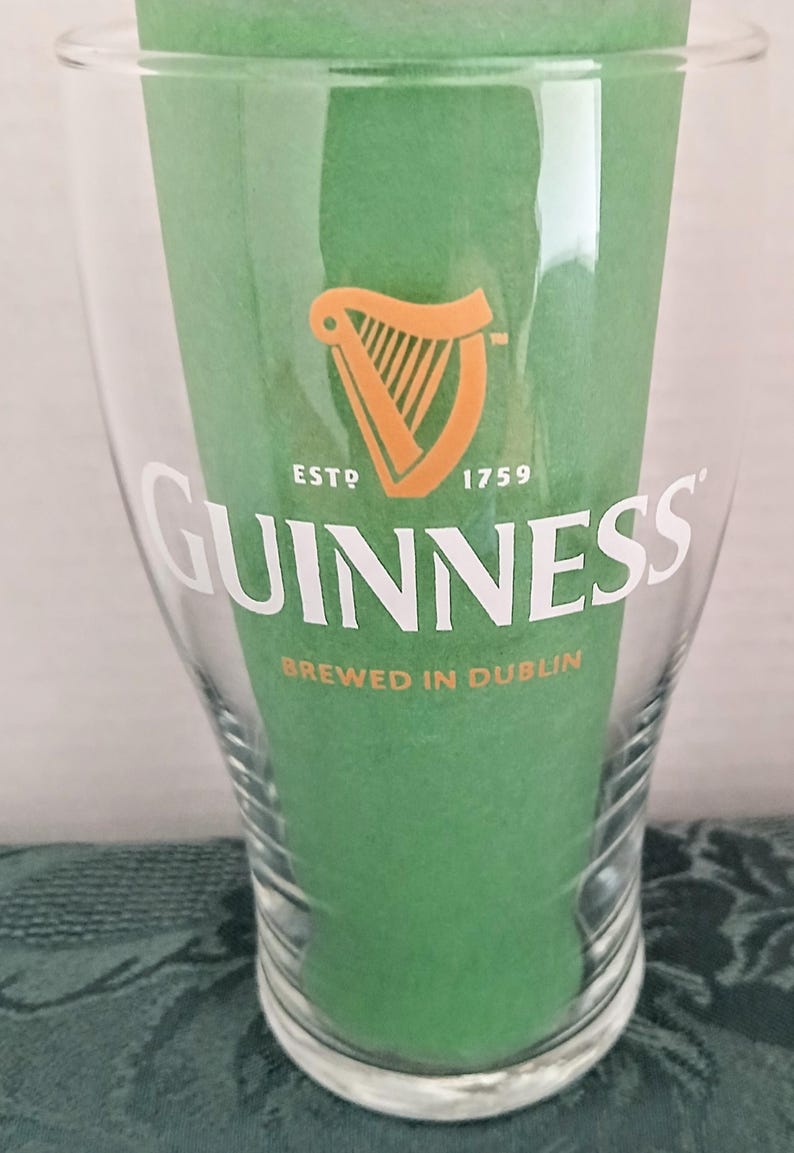 Może przedstawiać: Przezroczysty szklany kufel Guinness z białym logo Guinness i pomarańczowym symbolem harfy. Widoczny jest r&oacute;wnież tekst "ESTD 1759" i "BREWED IN DUBLIN". Szklanka wypełniona jest zielonym papierem.