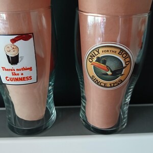 Może przedstawiać: Dwa przezroczyste szklanki do piwa z marką Guinness. Jedna szklanka ma etykietę z napisem "There's nothing like a Guinness" i obrazem nalewanego piwa. Druga szklanka ma etykietę z tukanem i napisem "Only for the Bold Since 1759."