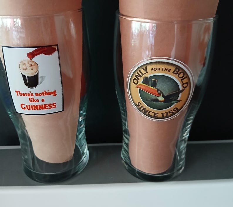 Może przedstawiać: Dwa przezroczyste szklanki do piwa z marką Guinness. Jedna szklanka ma etykietę z napisem "There's nothing like a Guinness" i obrazem nalewanego piwa. Druga szklanka ma etykietę z tukanem i napisem "Only for the Bold Since 1759."