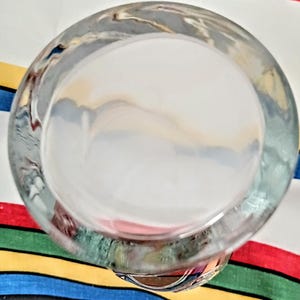 May include: A clear, round glass bowl with a thick rim. The bowl is positioned at an angle, offering a view into its interior. The background features a colorful striped fabric.