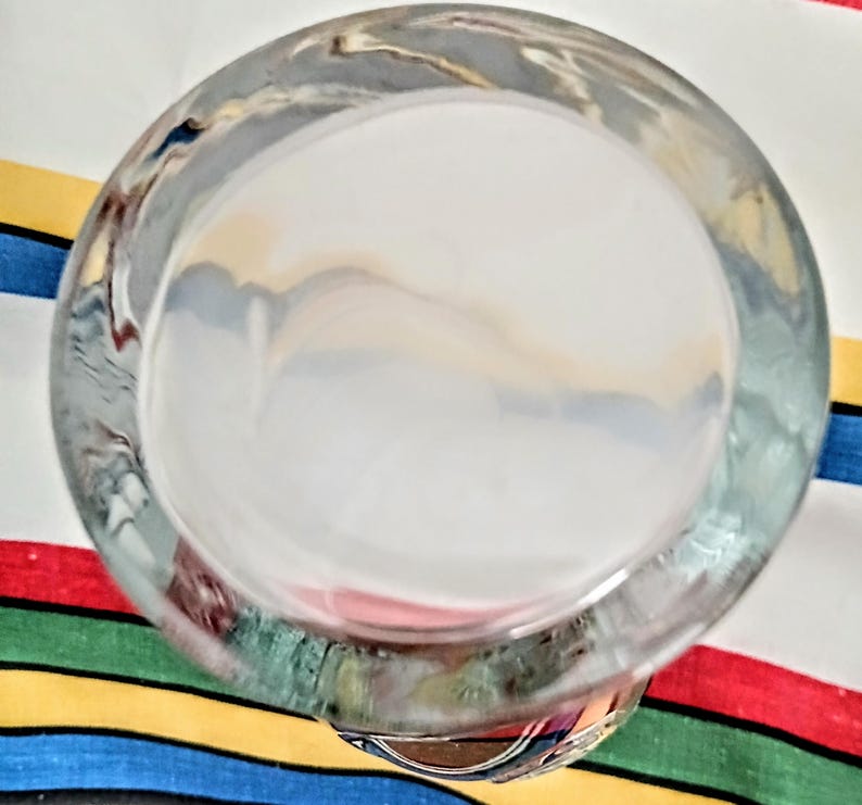 May include: A clear, round glass bowl with a thick rim. The bowl is positioned at an angle, offering a view into its interior. The background features a colorful striped fabric.