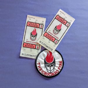 May include: Two rectangular paper tickets for a Chicago Fire versus Honolulu Hawaiians game on October 23, 1974, at Soldier Field, priced at $7.00, alongside a circular embroidered patch featuring the team logo with a red flame and black helmet.