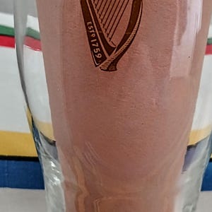 May include: A clear pint glass with the Guinness logo, a harp, and the text "ESTD 1759". The glass is partially filled with a brown liquid, and the background is blurred.