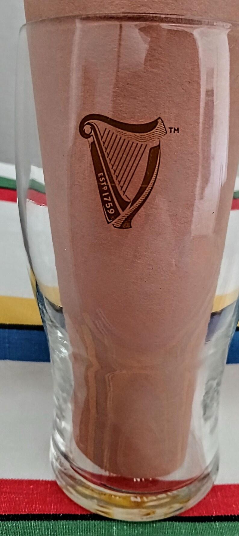 May include: A clear pint glass with the Guinness logo, a harp, and the text "ESTD 1759". The glass is partially filled with a brown liquid, and the background is blurred.