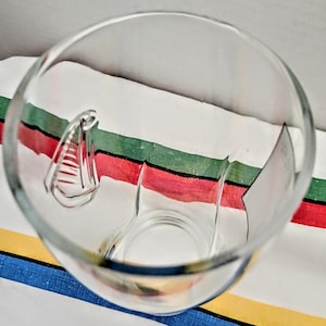 May include: A clear glass with a unique handle design. The glass is empty and sits on a striped fabric with red, green, and white stripes. The glass is transparent, allowing a view through its interior.