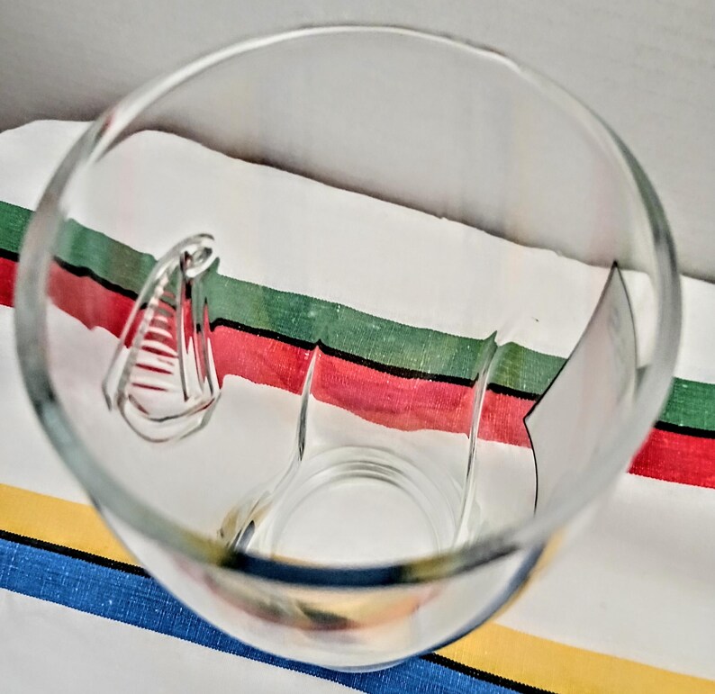 May include: A clear glass with a unique handle design. The glass is empty and sits on a striped fabric with red, green, and white stripes. The glass is transparent, allowing a view through its interior.