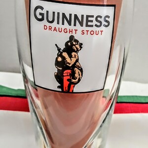 May include: A clear glass with a unique, twisted design. The glass features the Guinness logo in white with red text, and an illustration of a bear carrying a keg. The glass is designed for draught stout.