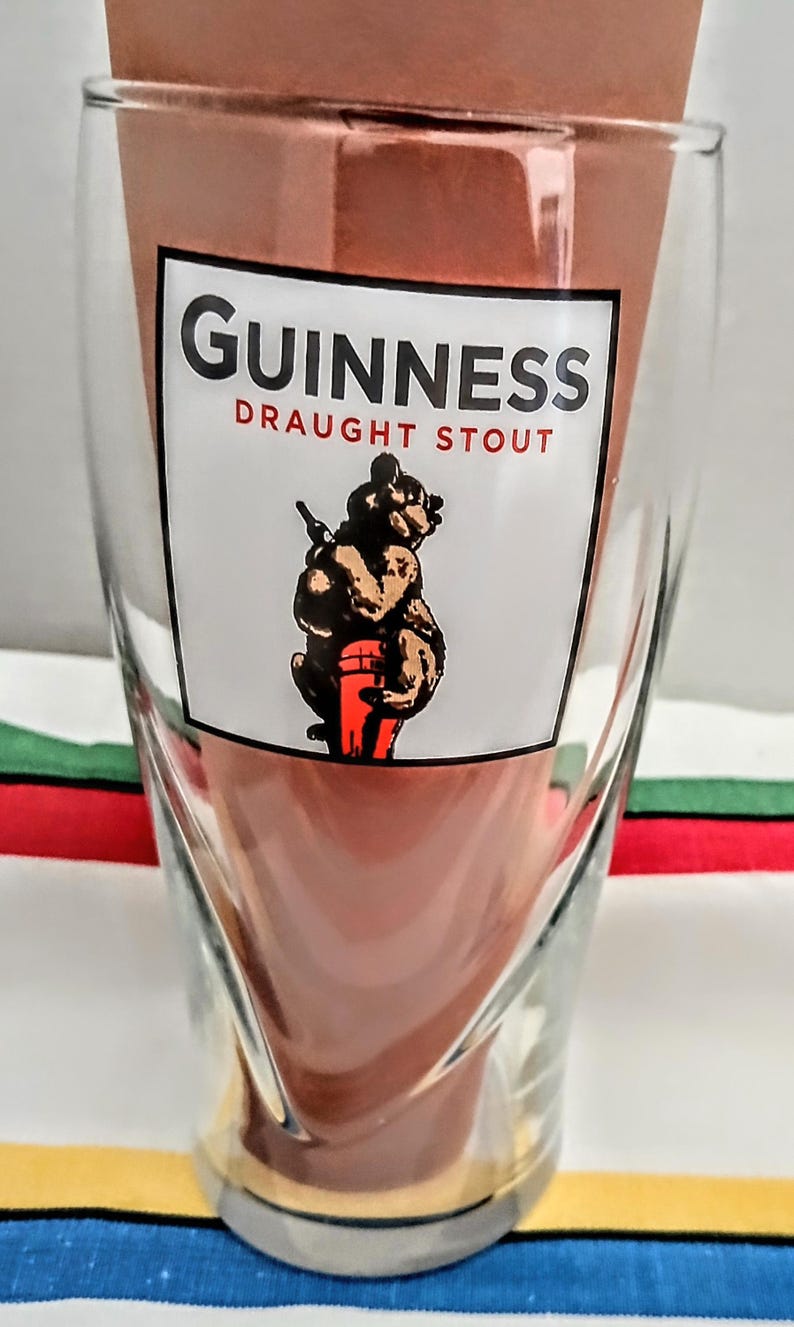 May include: A clear glass with a unique, twisted design. The glass features the Guinness logo in white with red text, and an illustration of a bear carrying a keg. The glass is designed for draught stout.