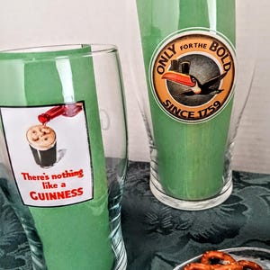 Może przedstawiać: Dwa przezroczyste szklanki do piwa z zielonymi papierowymi wkładkami. Jedna szklanka ma napis "There's nothing like a Guinness" i obraz wlewanego Guinnessa. Druga szklanka ma okrągłe logo z tukanem i napisem "Only for the Bold Since 1759".