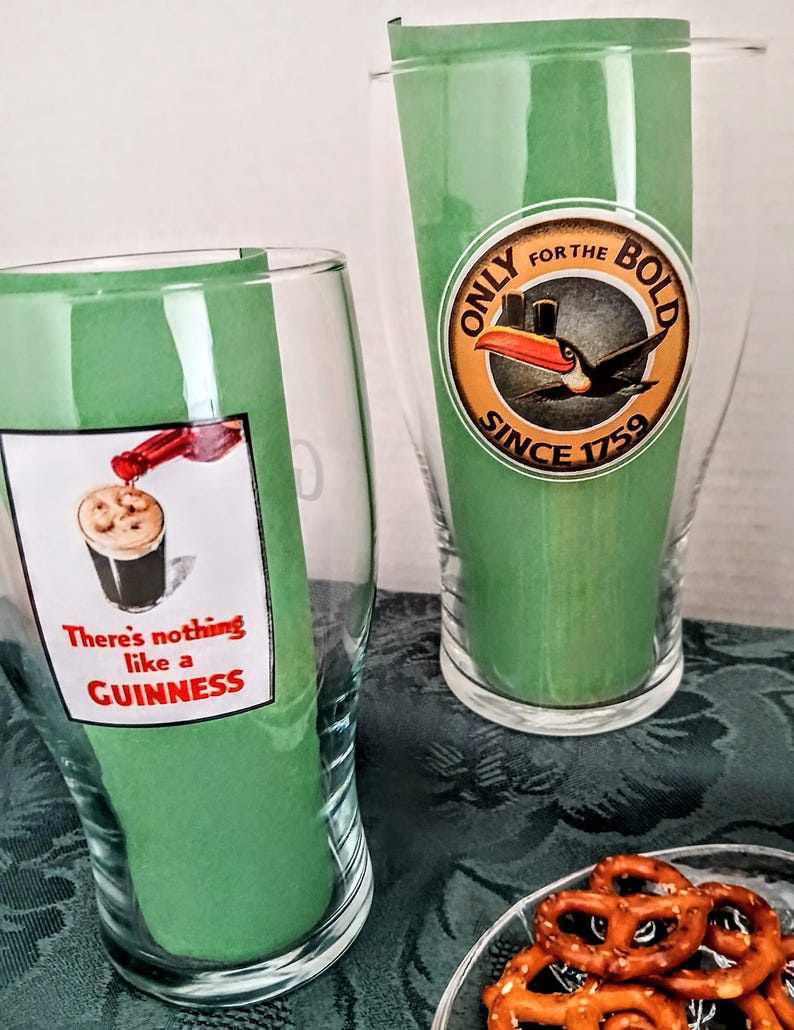 Może przedstawiać: Dwa przezroczyste szklanki do piwa z zielonymi papierowymi wkładkami. Jedna szklanka ma napis "There's nothing like a Guinness" i obraz wlewanego Guinnessa. Druga szklanka ma okrągłe logo z tukanem i napisem "Only for the Bold Since 1759".