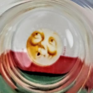 May include: A clear glass object with a circular design featuring a yellow and brown face. The face is centered within the glass, and the background includes red, white, and green colors.