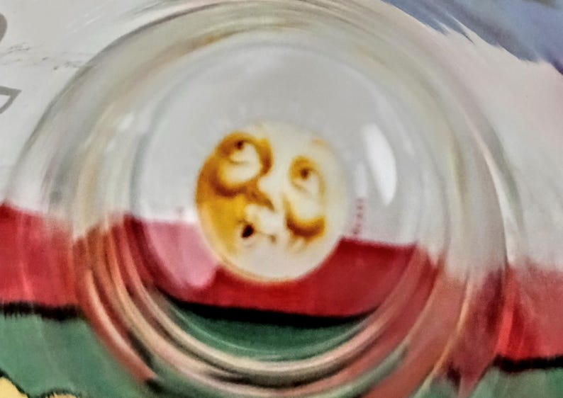 May include: A clear glass object with a circular design featuring a yellow and brown face. The face is centered within the glass, and the background includes red, white, and green colors.