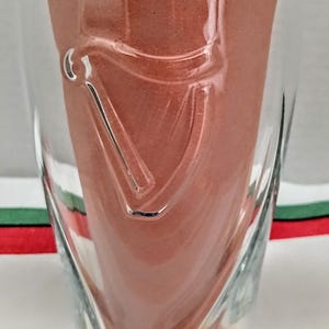 May include: A clear glass with a curved design and a decorative emblem. The glass is filled with a reddish-brown material. The glass sits on a striped surface with blue, yellow, green, and red stripes.