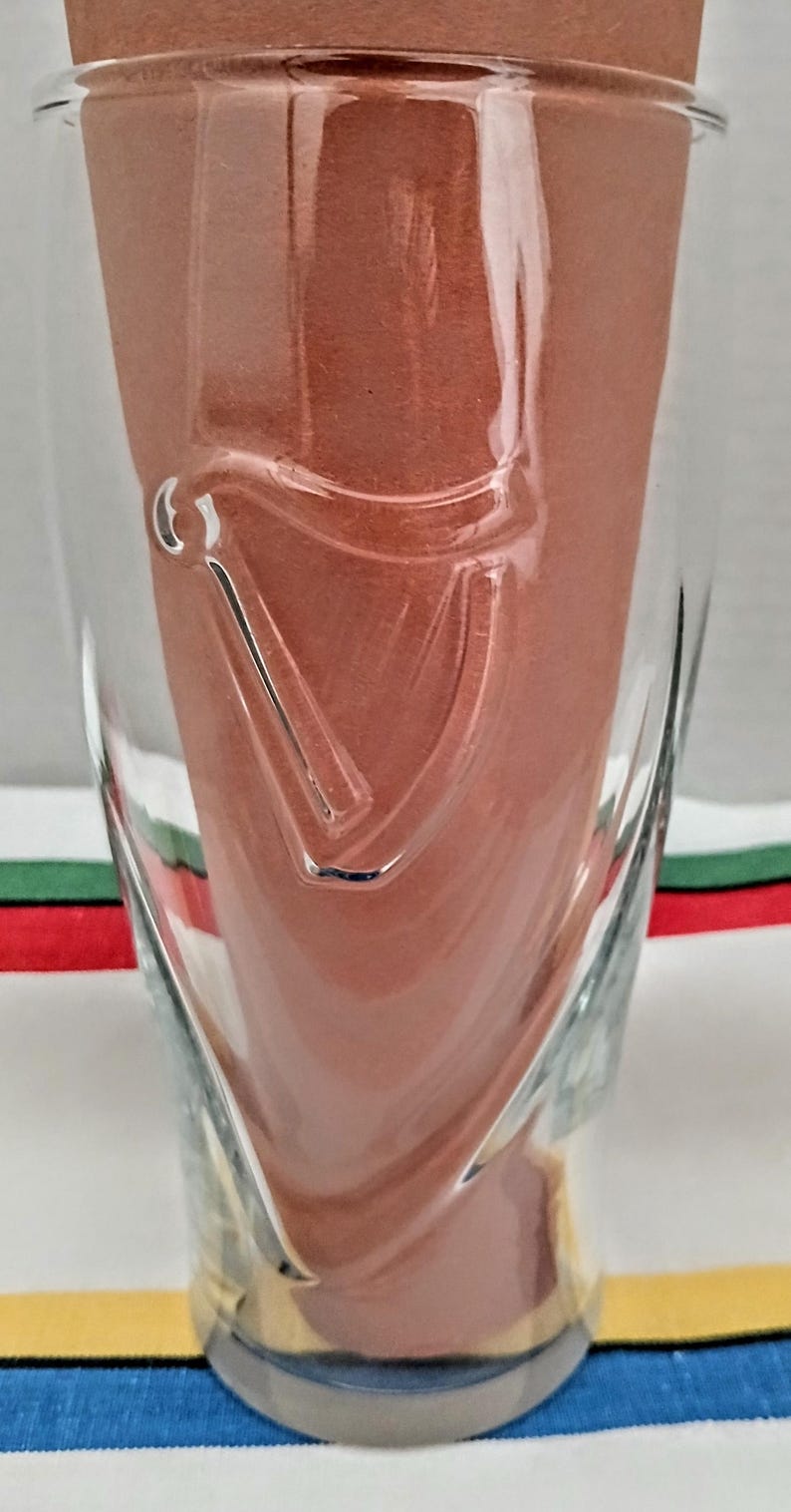 May include: A clear glass with a curved design and a decorative emblem. The glass is filled with a reddish-brown material. The glass sits on a striped surface with blue, yellow, green, and red stripes.