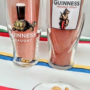 May include: Two clear Guinness pint glasses with unique designs. One features a turtle with a pint of stout, the other a bear with a pint. The glasses have the Guinness logo and are set against a striped background.
