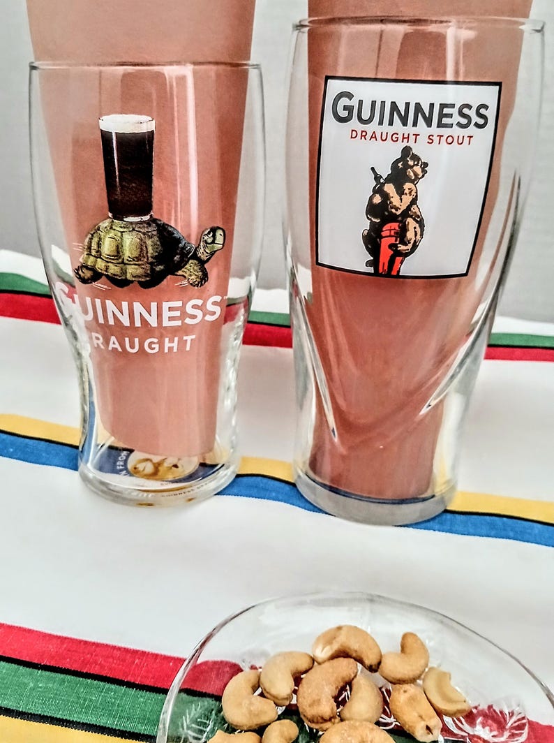 May include: Two clear Guinness pint glasses with unique designs. One features a turtle with a pint of stout, the other a bear with a pint. The glasses have the Guinness logo and are set against a striped background.