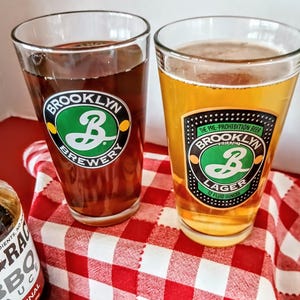 May include: Two pint glasses filled with beer, one dark and one light, sit on a red and white checkered cloth. The glasses feature the "Brooklyn Brewery" logo. One glass is labeled "Brooklyn Lager, 12 fluid ounces."