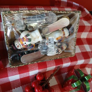 May include: A rectangular tray filled with various snowman-themed spreaders and knives, wrapped in clear plastic. The tray sits on a red and white checkered tablecloth, with a sprig of red berries and a small gift box with a green ribbon.