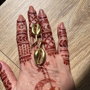 May include: Gold ring with two shell-shaped charms. The ring is worn on the middle finger of a hand with henna.