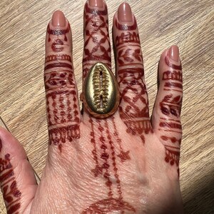 May include: A gold ring with a shell design. The ring is worn on the middle finger of a hand with henna tattoos.
