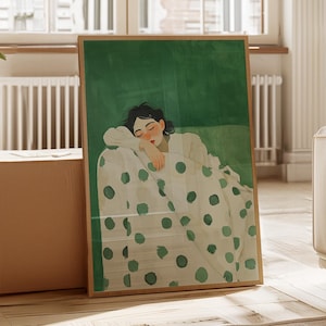 May include: A framed art print depicting a woman resting on a polka-dot patterned surface against a green background. The artwork is in a wooden frame and is displayed in a bright room with a large window and a potted plant.