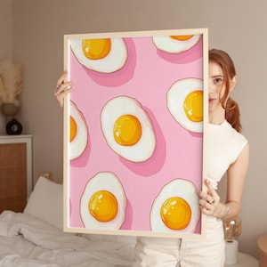 Può includere: Stampa incorniciata con un motivo ripetuto di uova fritte in stile cartone animato su sfondo rosa. Ogni uovo ha un tuorlo giallo brillante e un albume bianco. La stampa è in una cornice di legno chiaro.