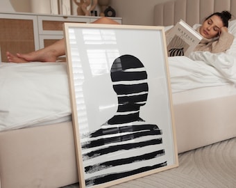 May include: A black and white abstract art print of a person's silhouette with horizontal brushstrokes. The print is framed in a natural wood frame and is leaning against a white bed.