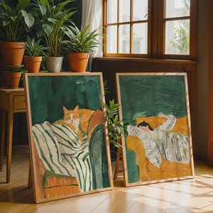 Pode incluir: Duas impressões artísticas emolduradas. Uma retrata um gato laranja descansando em uma cadeira, coberto por um cobertor listrado. A outra mostra uma pessoa dormindo em um sofá amarelo, também coberta com um cobertor listrado. Ambas estão em molduras de madeira.