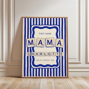 May include: Framed art print with a blue and white striped background. The print features the word "MAMA" spelled out in Scrabble tiles, with the name "CHARLOTTE" below. Text at the top reads "FIRST WORD" and at the bottom "SAID AT 11 MONTHS - 2026".