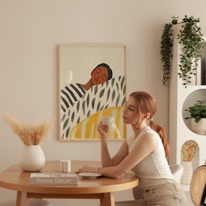 Op de afbeelding: Een vrouw zit aan een tafel met een witte mok in haar hand en kijkt naar de mok. Ze zit voor een ingelijst afdruk van een vrouw met gesloten ogen, liggend op een bed van geel, groen en zwart gepatroneerd textiel. De afdruk is op een witte muur. De tafel is van hout en heeft een boek erop dat "Home Decor" zegt.