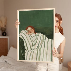 Può includere: Una stampa artistica incorniciata raffigurante una donna addormentata, avvolta in una coperta a righe. L'opera d'arte ha uno sfondo verde e una cornice in legno chiaro. La donna ha i capelli rossi e appoggia la testa sul braccio.