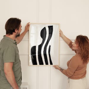 Może przedstawiać: Dwie osoby wieszają na białej ścianie oprawiony czarno-biały wydruk abstrakcyjnego dzieła sztuki. Wydruk przedstawia stylizowaną czarną postać z białymi liniami.