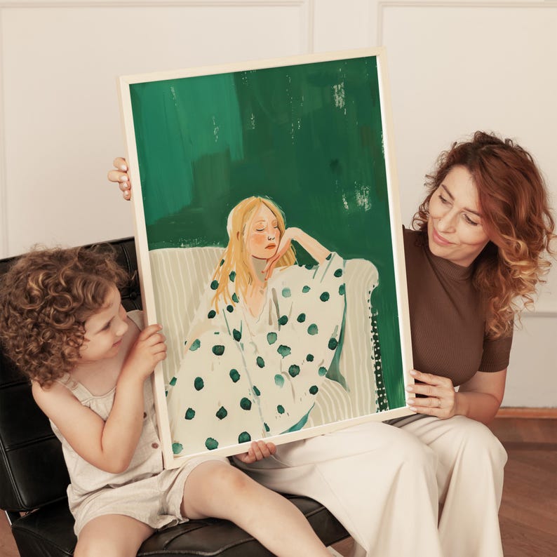 May include: A framed art print featuring a woman with blonde hair in a white robe with green polka dots, resting on a sofa. The background is a textured green. The print is held by a woman and a child.
